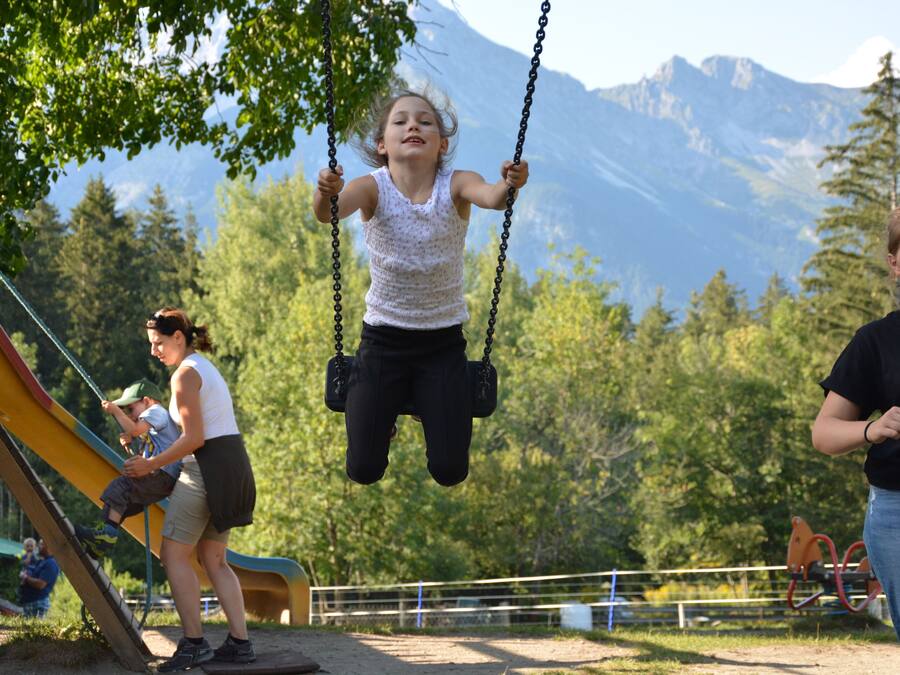 A playground with swings and a slide, surrounded by trees and mountains in the background.