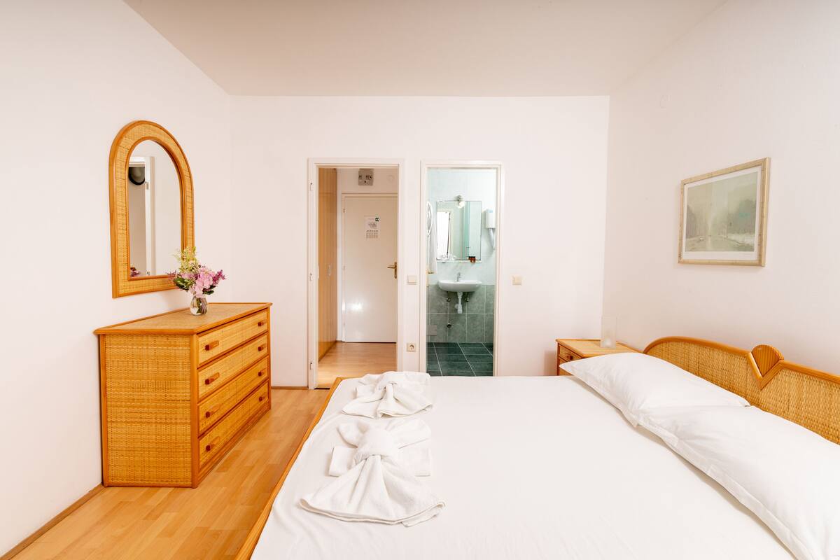 A bedroom with a wooden dresser, a large bed with white linens, a mirror, and a framed picture on the wall.