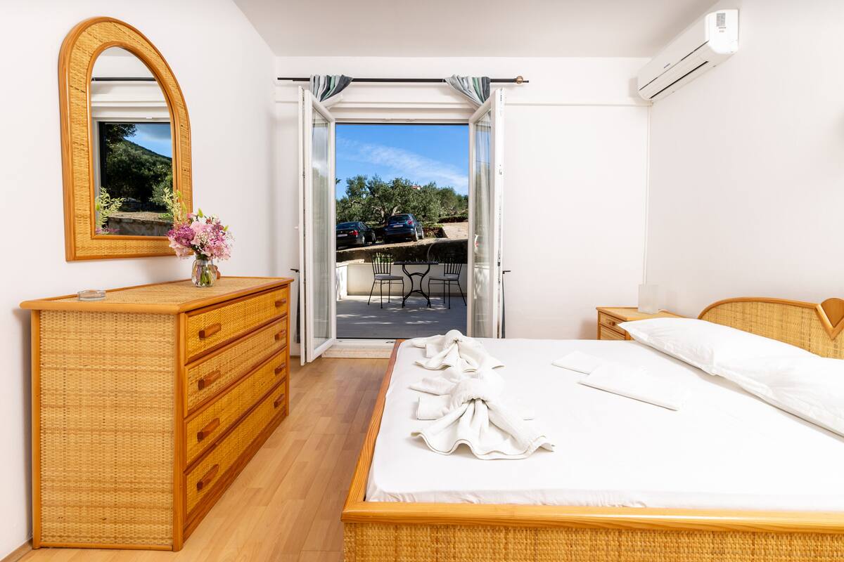 A bedroom with a bed, a dresser, a mirror, and an open door leading to a balcony with a view of parked cars and greenery.