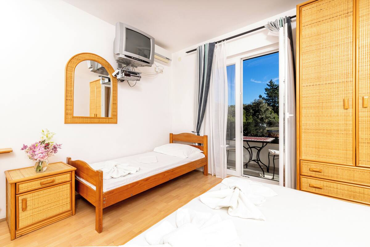 A bedroom with a wooden bed, a bedside table with a vase of flowers, a television mounted on the wall, and a balcony with a table and chairs.