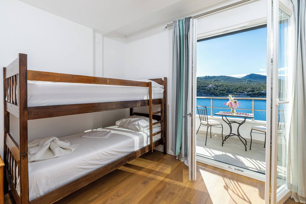 A room with a bunk bed, a balcony with a table and chairs, and a view of the sea and hills.