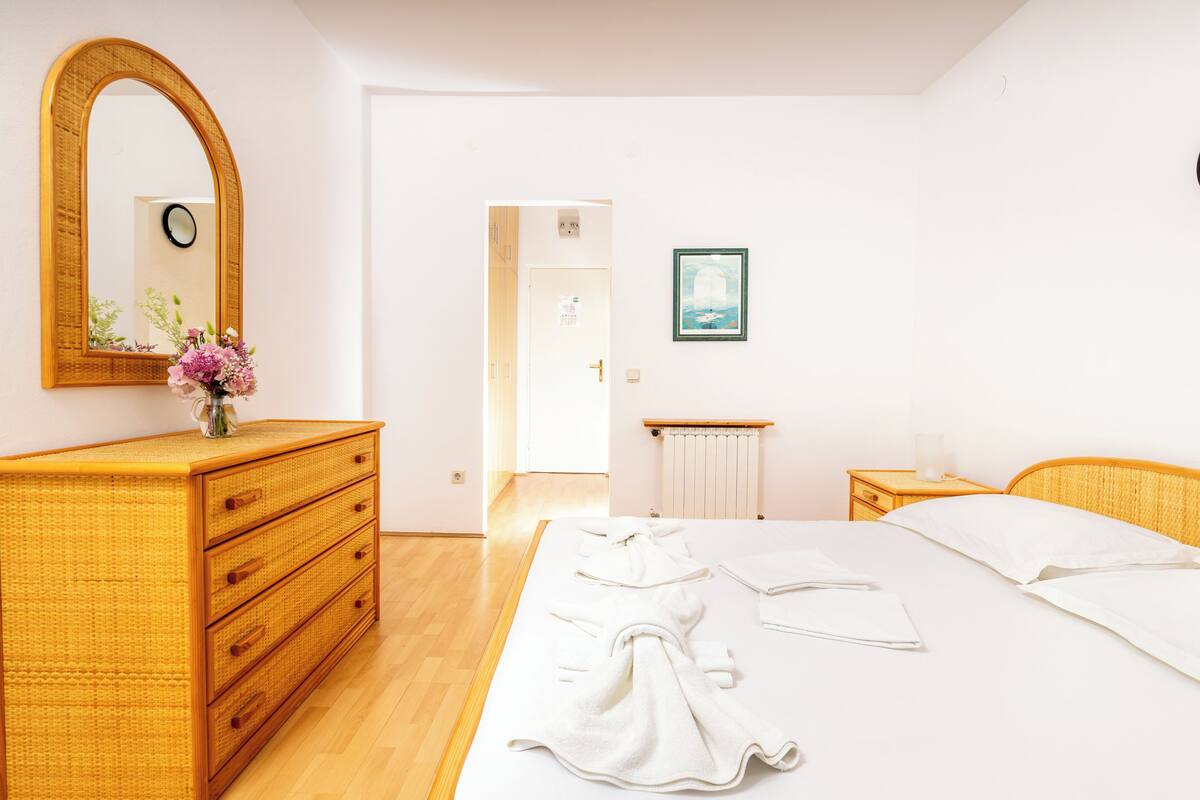 A hotel room with a wooden dresser, a bed with white linens, a mirror, and a framed picture on the wall.
