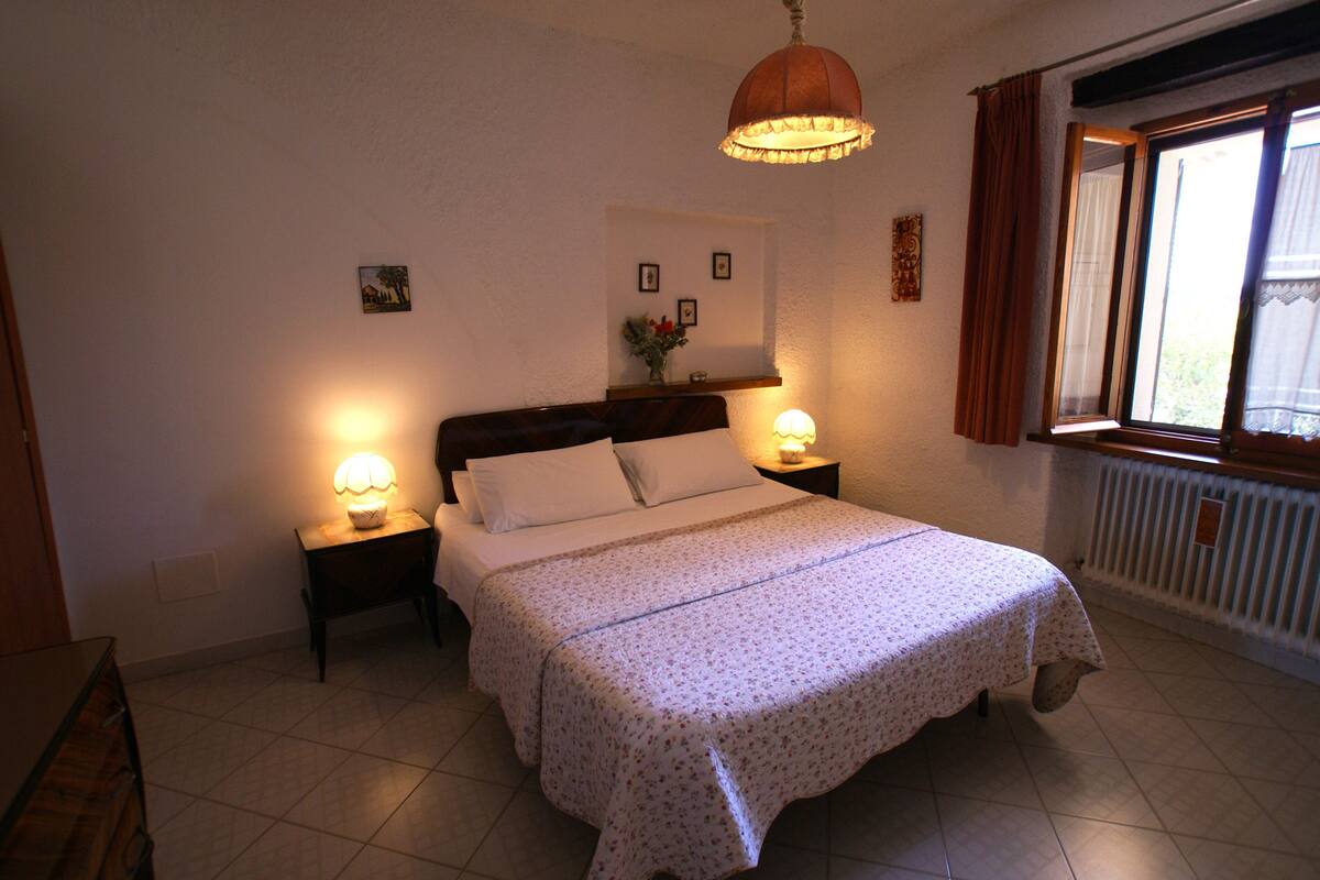 Een slaapkamer met een bed, twee bedlampen, een nachtkastje, een raam met gordijnen en een wandplank met foto's.