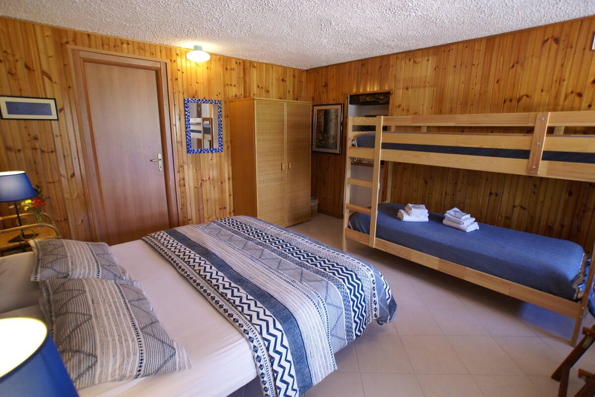 Een kamer met houten wanden, een stapelbed, een enkel bed met beddengoed met dessin, een kledingkast en een bureau met een lamp.