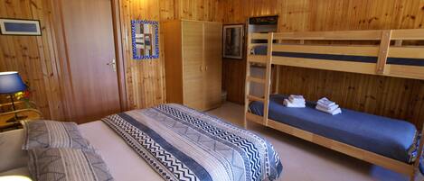 A room with wooden walls, a bunk bed, a single bed with patterned bedding, a wardrobe, and a desk with a lamp.