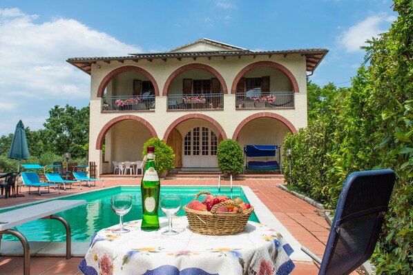 Un espace piscine avec une table dressée pour deux personnes, une bouteille de vin et un panier de fruits.