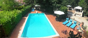 Une piscine rectangulaire, bordée d’une terrasse carrelée, entourée de verdure et ombragée par des arbres.