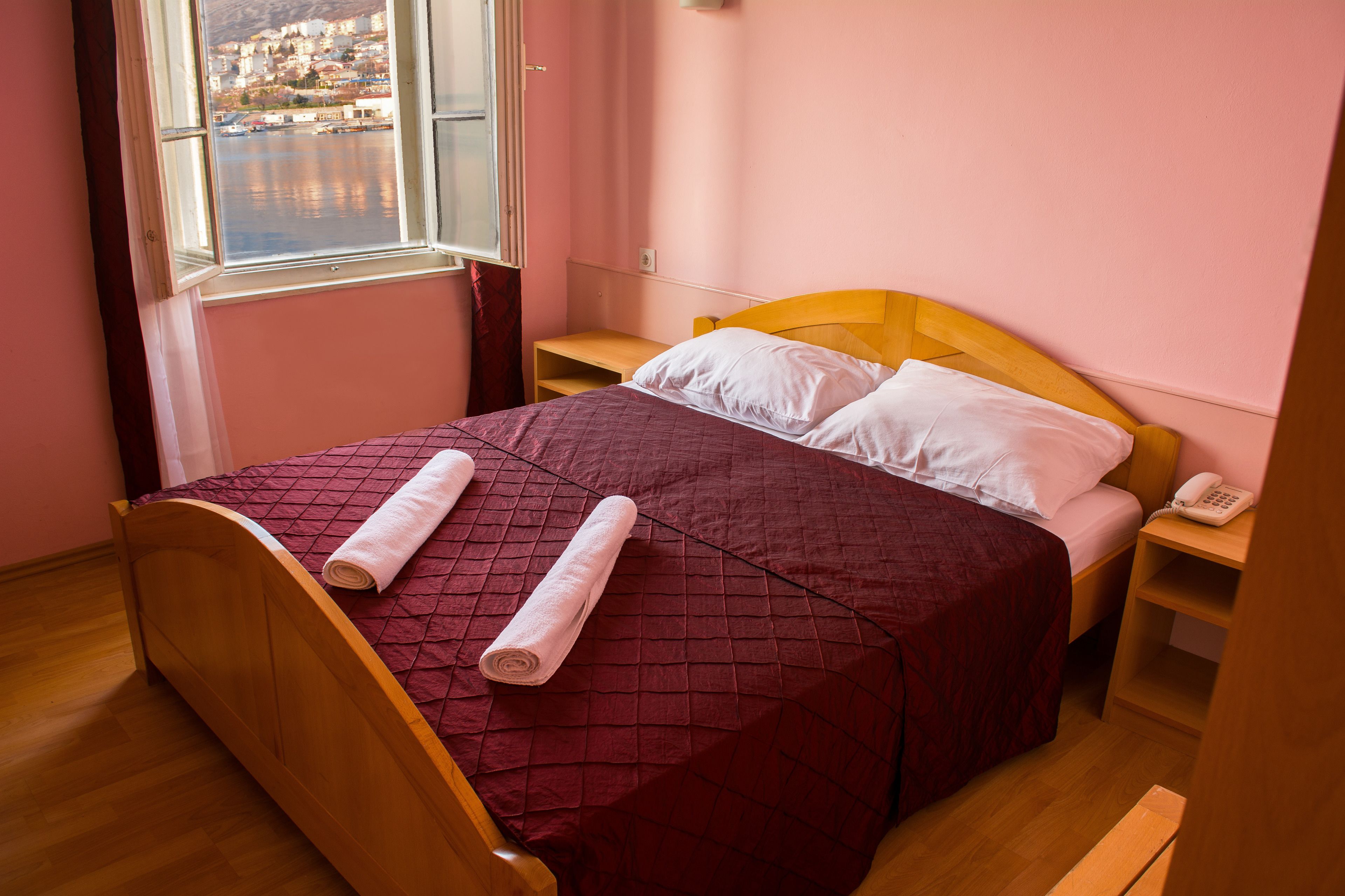 Ein ordentlich bezogenes Bett mit einer roten Steude, einem hölzernen Kopfteil und Blick auf ein Gewässer durch ein Fenster.