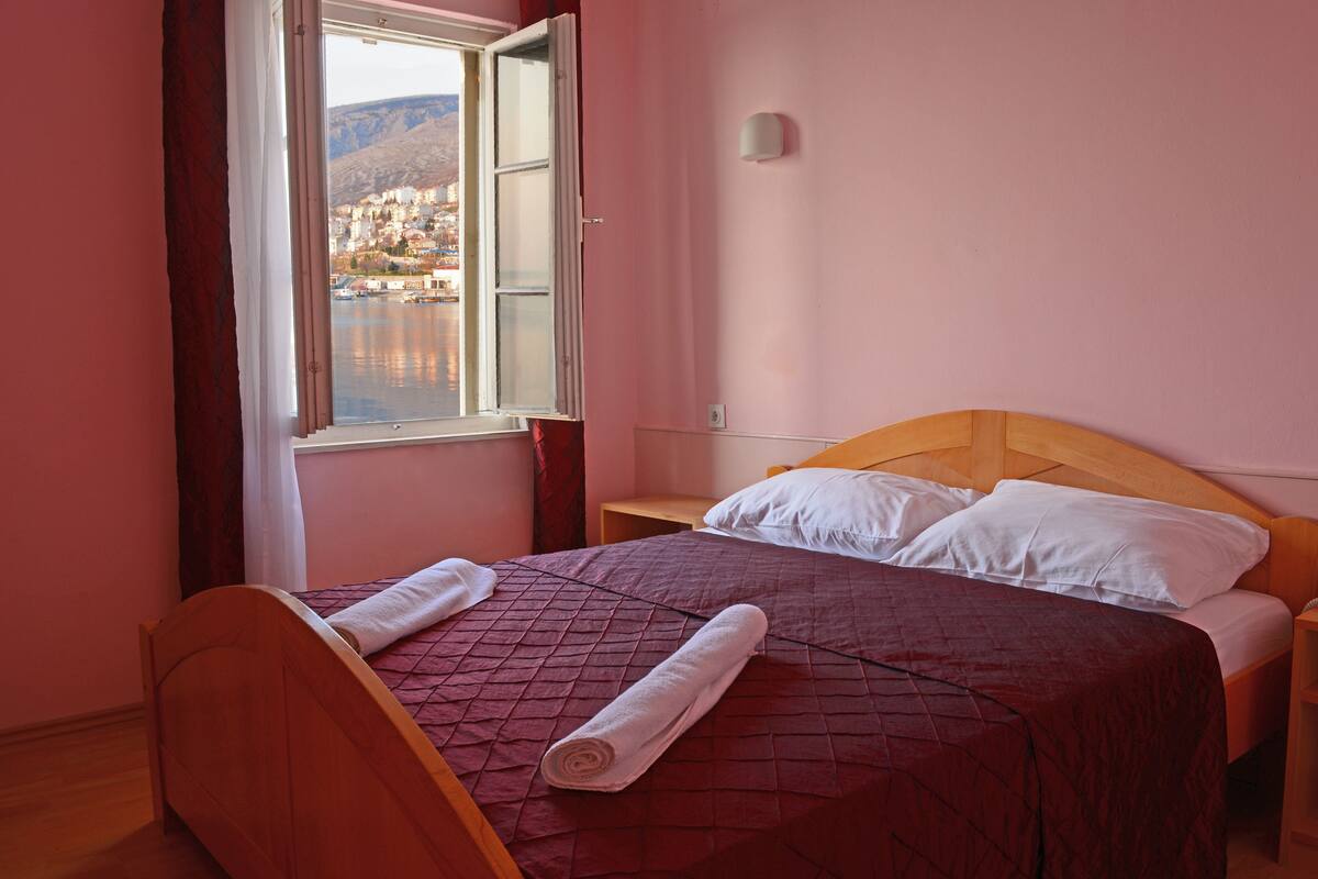 A room with a bed, a nightstand, a window with curtains, and a view of a town.