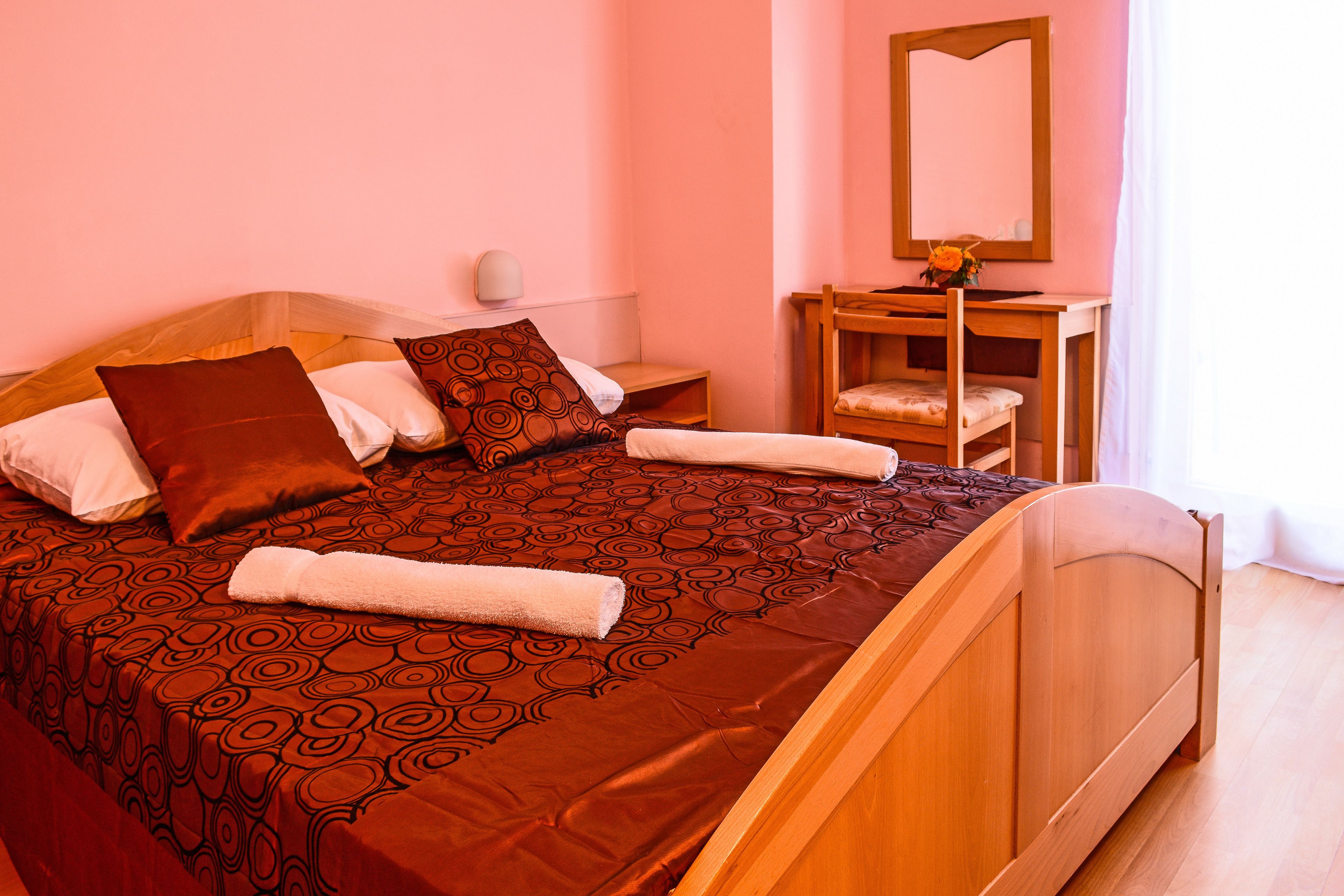 Een netjes opgemaakt bed met een bedsprei met patroon, een houten nachtkastje met een lamp en een stoel met een kussen.