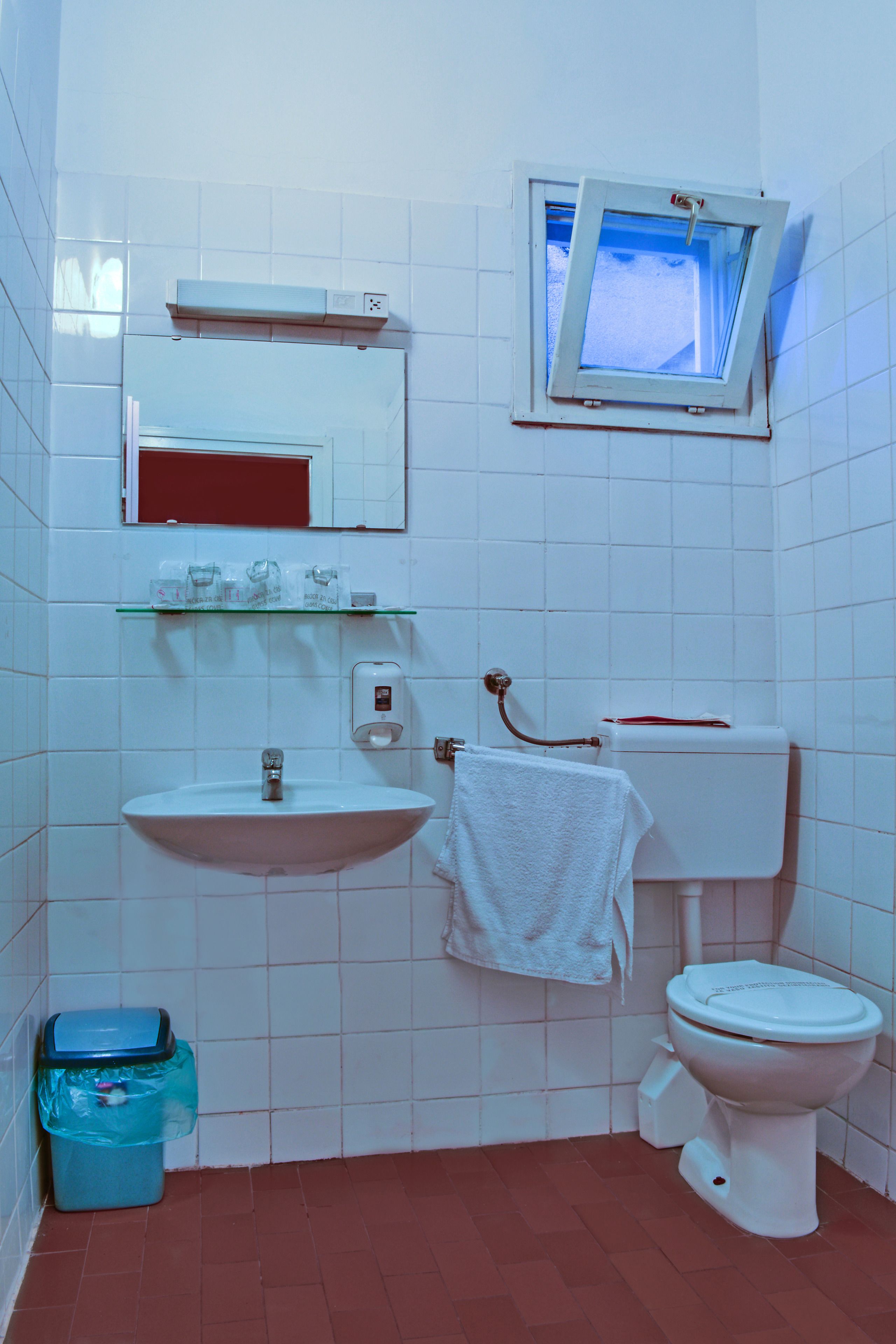 Ein Badezimmer mit Waschbecken, Toilette, Fenster und einer blauen Mülltonne.