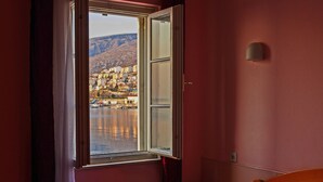 A hotel room with a bed, a nightstand, a window with a view of a cityscape, and a wall-mounted light fixture. - Hotel Art (Senj)