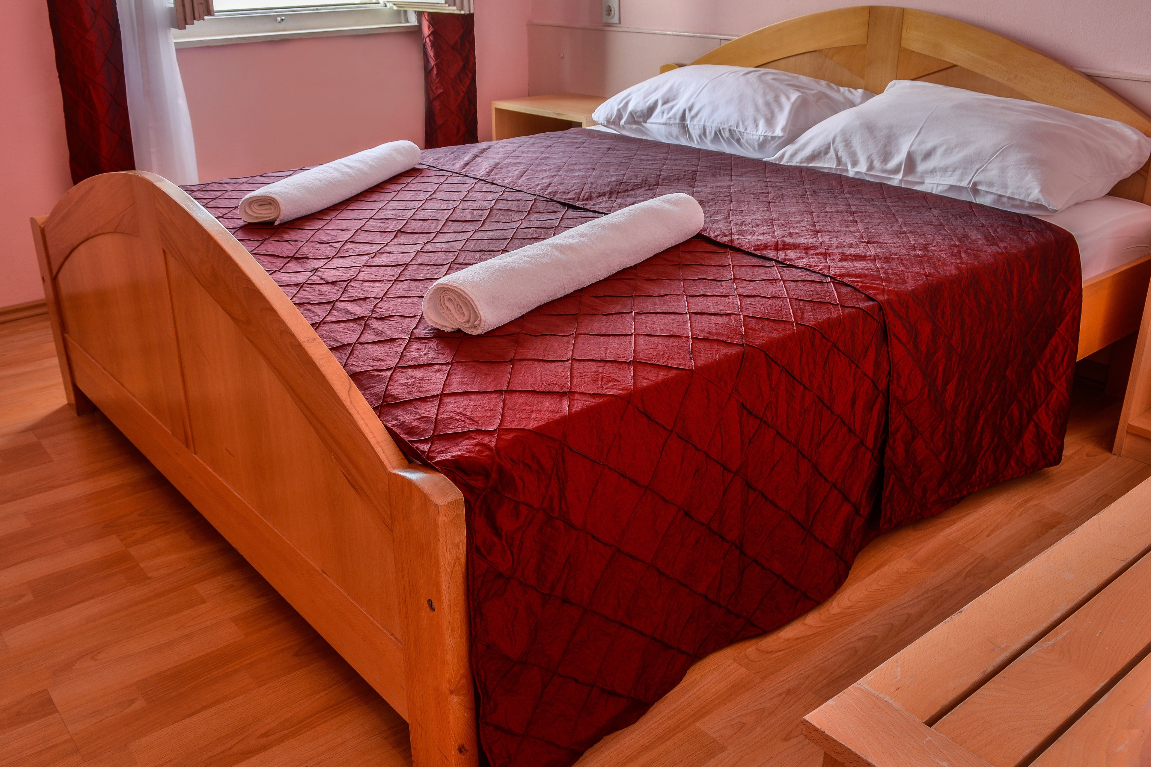 A neatly made bed with a red quilt, wooden headboard, and two rolled towels.