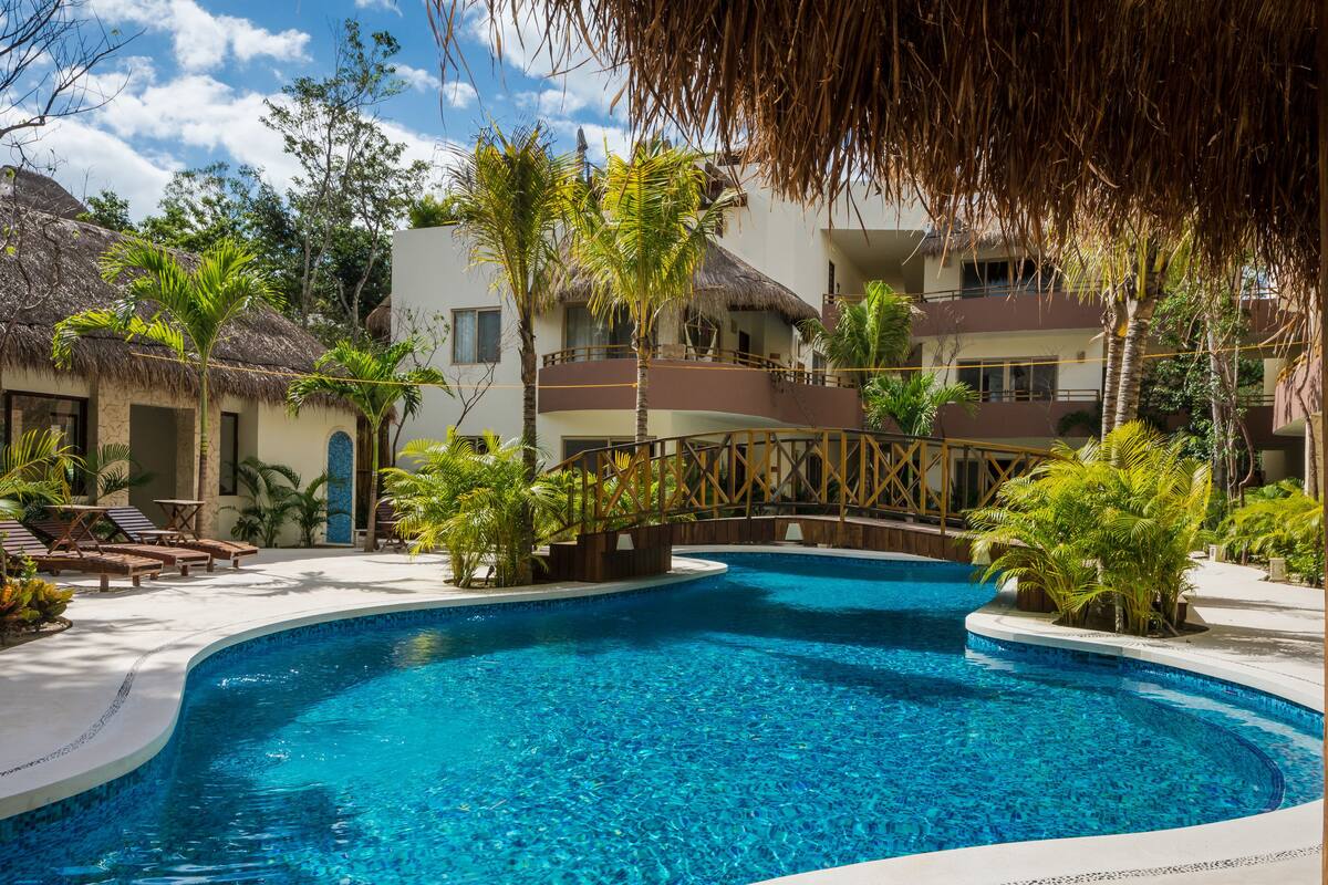 A swimming pool surrounded by lounge chairs, palm trees, and a building with a thatched roof.