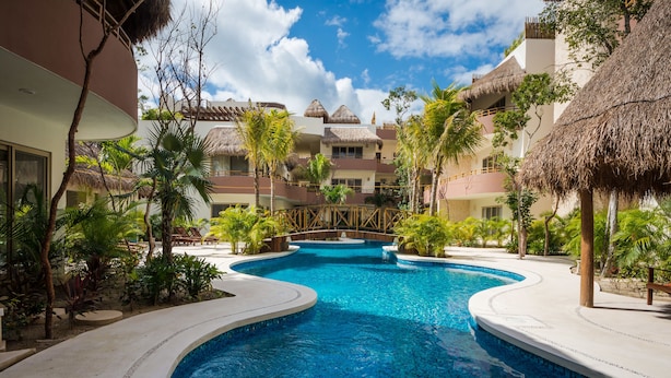 Ein Poolbereich eines Resorts mit zentralem Pool, Palmen und Gebäuden mit Reetdächern.