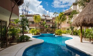 A resort pool area with a central pool, palm trees, and thatched-roof buildings. - Tulum Aqua Condos (Tulum)