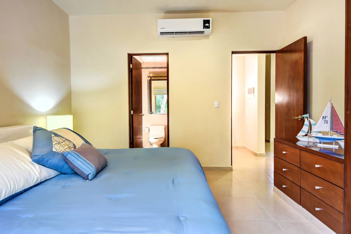 A bedroom with a bed, pillows, a dresser, a small sailboat model, and an air conditioning unit.