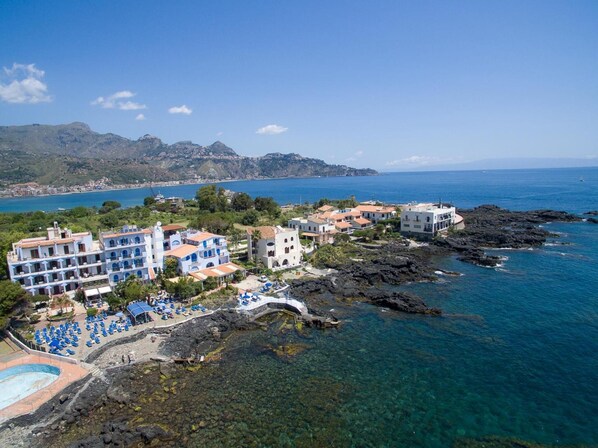 A coastal hotel with multiple buildings, a beach area with sun loungers, and clear blue waters. - Hotel Kalos (Giardini Naxos)