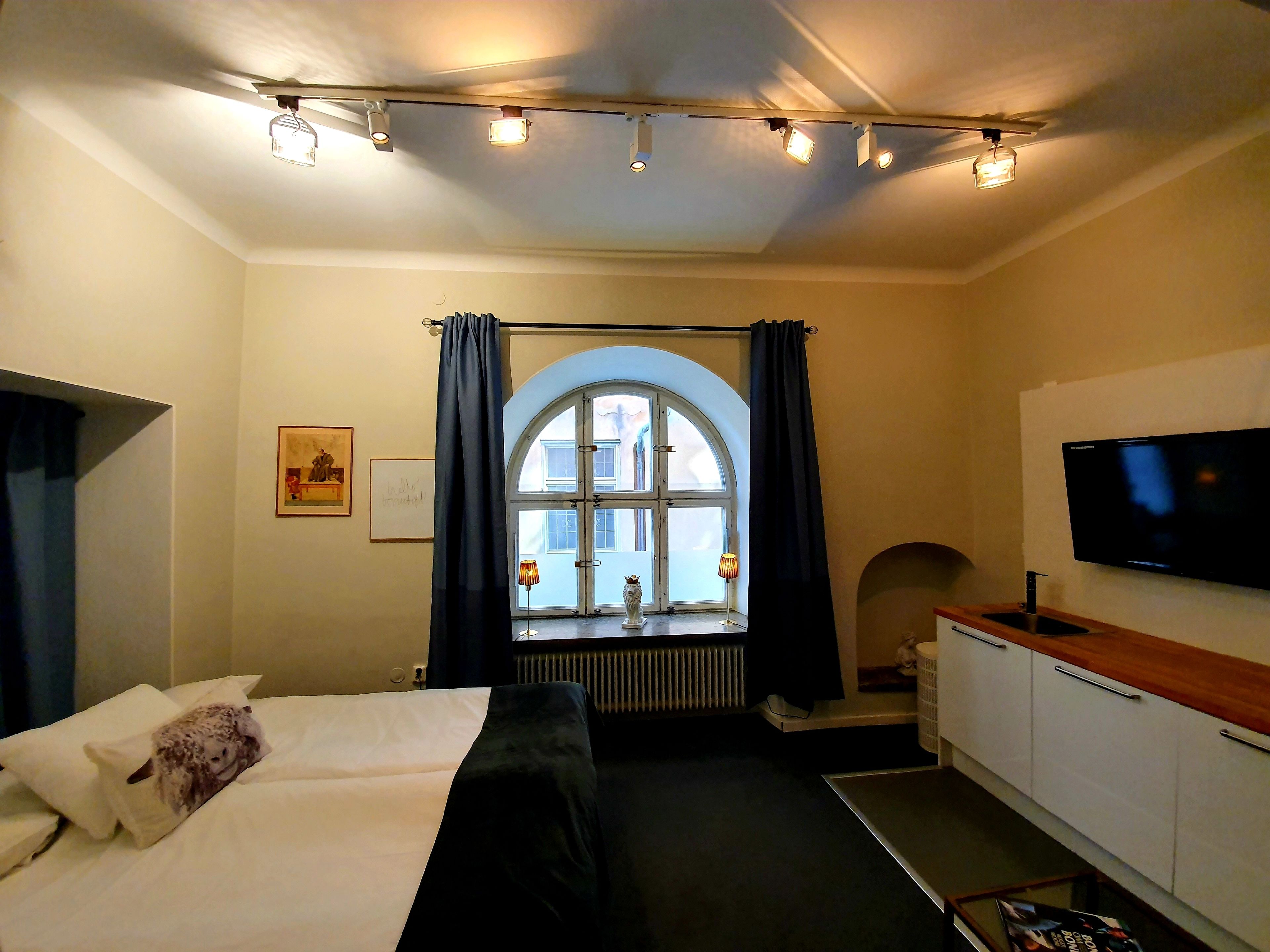 Quarto de hotel com cama, televisão, janela com cortinas, radiador e armário.