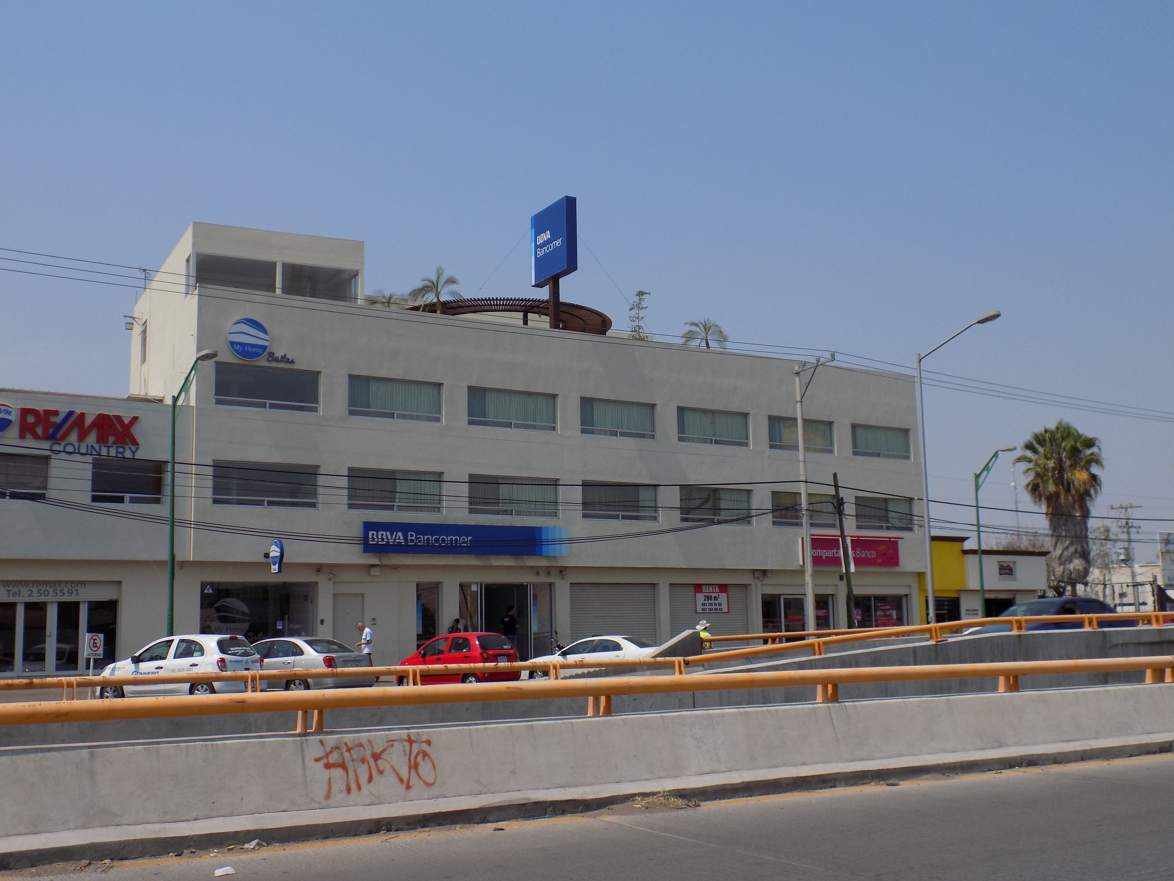 Un edificio de varios pisos con un letrero azul de BBVA Bancomer, un coche blanco estacionado al frente y un letrero visible de RE/MAX.