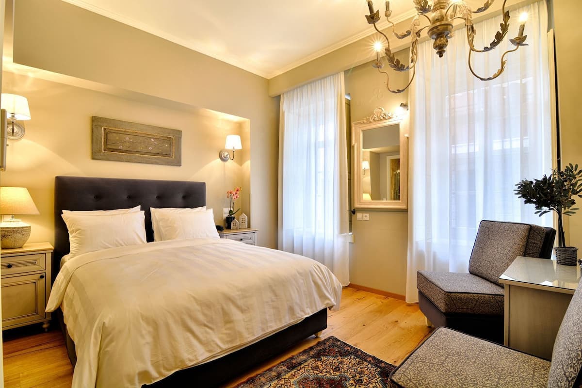 A hotel room with a large bed, a nightstand with a lamp, a chair, a table, and a chandelier.