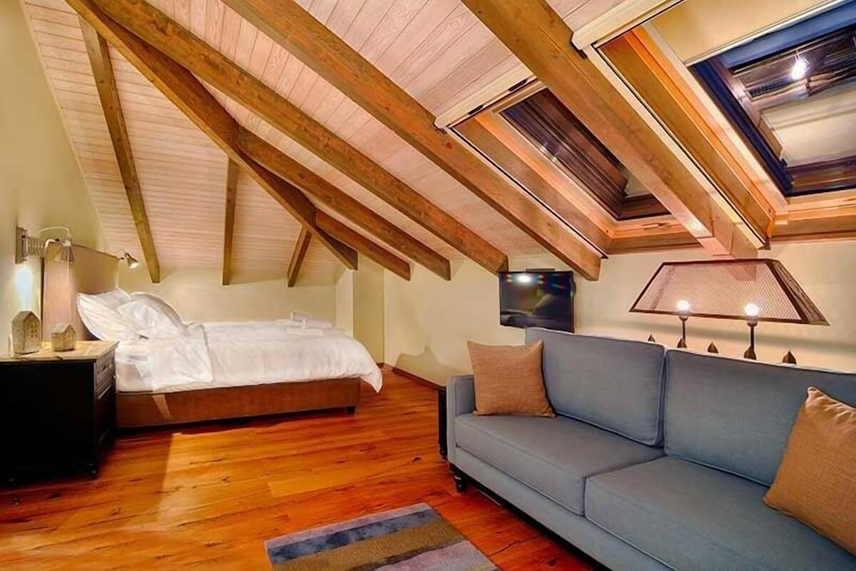 A bedroom with a sloped ceiling, wooden beams, a bed, a sofa, and a wall-mounted TV.