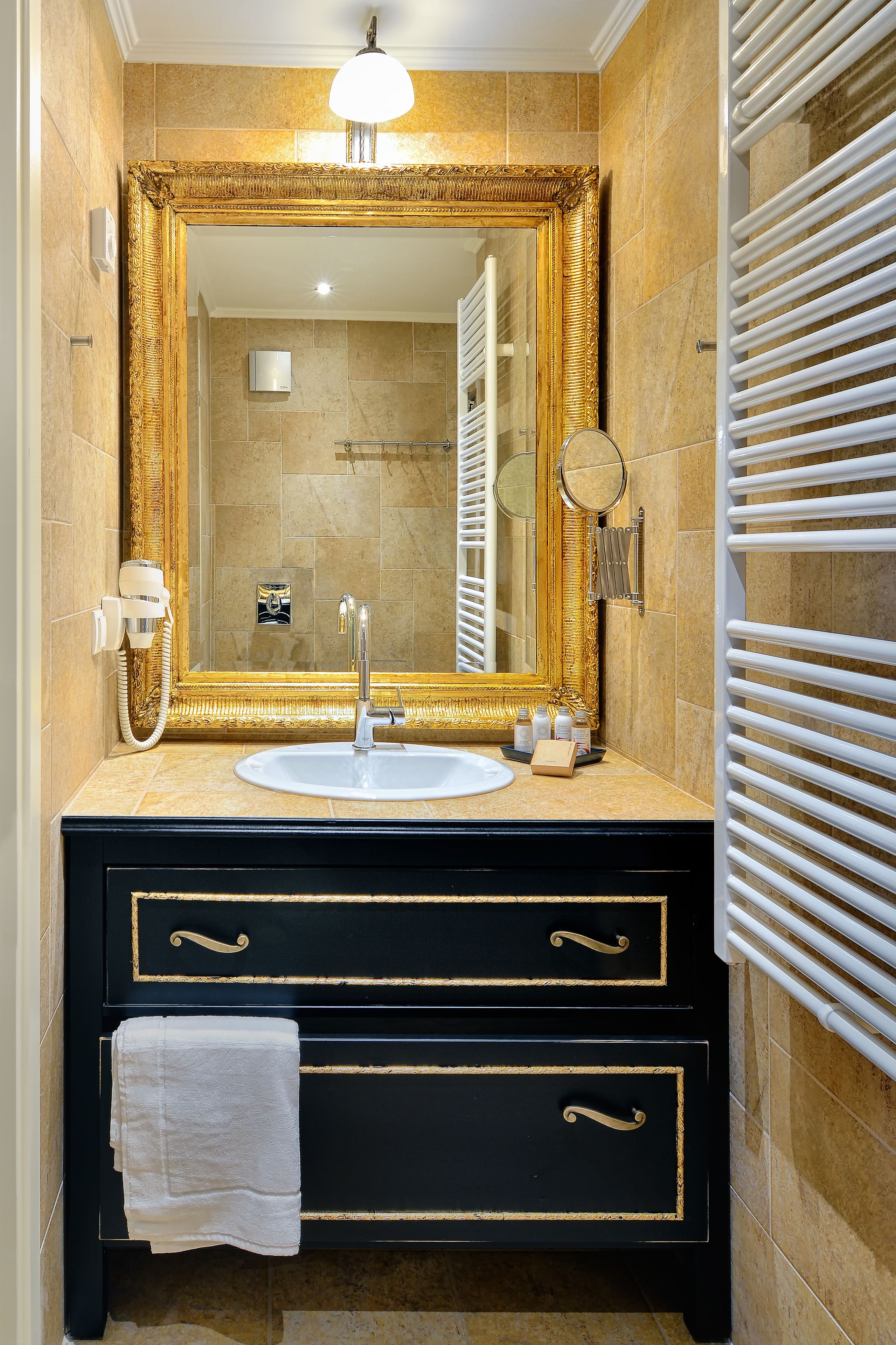 Une salle de bain avec un miroir encadré d’or, un lavabo blanc et un meuble-lavabo noir orné de motifs dorés.