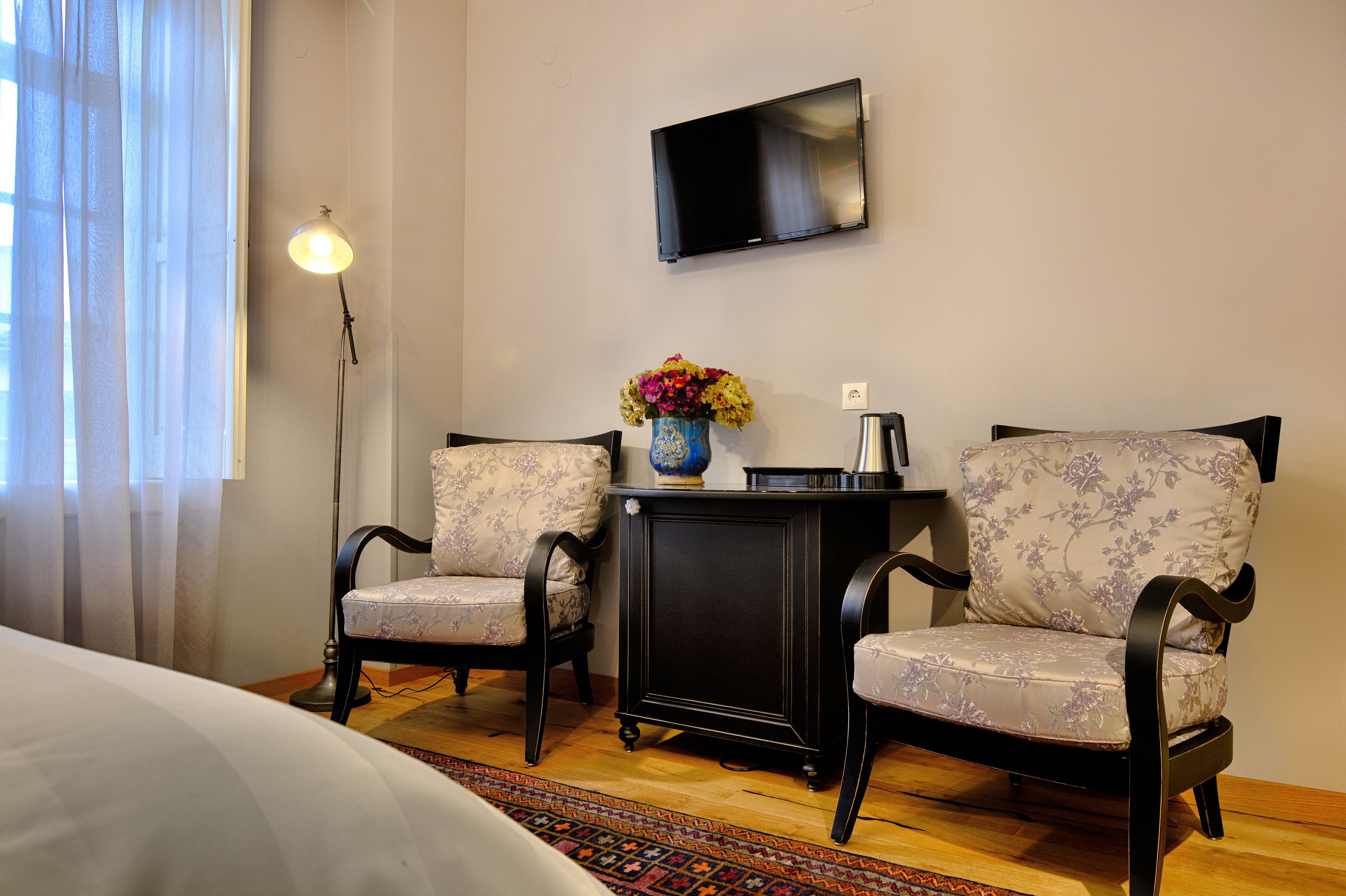 A hotel room with two upholstered chairs, a small table with a vase of flowers, a flat-screen TV mounted on the wall, and a floor lamp.