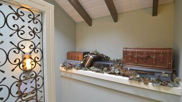 Ein Zimmer mit weißer Decke, Holzbalken, einem dekorativen Metallgitter und einem Regal mit alten Koffer und Pflanzen.