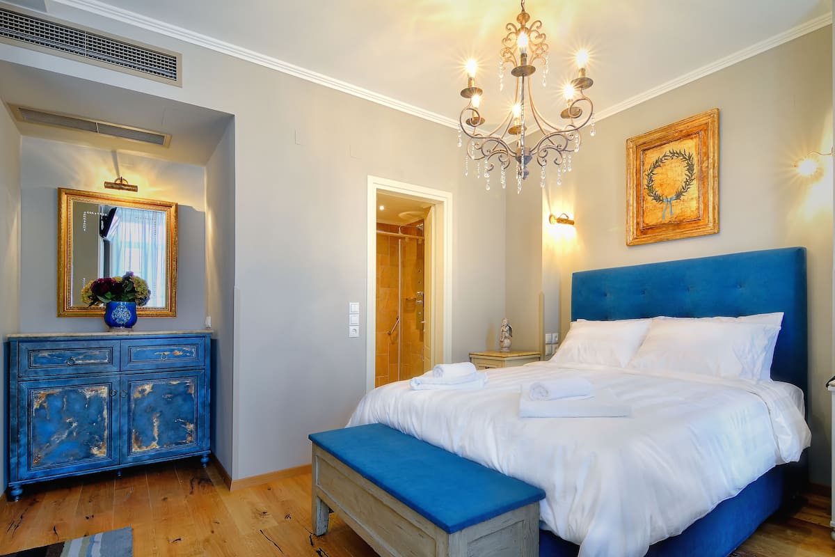 A bedroom with a large bed, a blue bench, a dresser, a mirror, a chandelier, and a door leading to a bathroom.