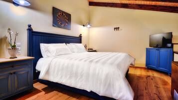A bedroom with a wooden floor, a bed with white bedding, a nightstand with a lamp, a dresser, and a television on a blue cabinet.