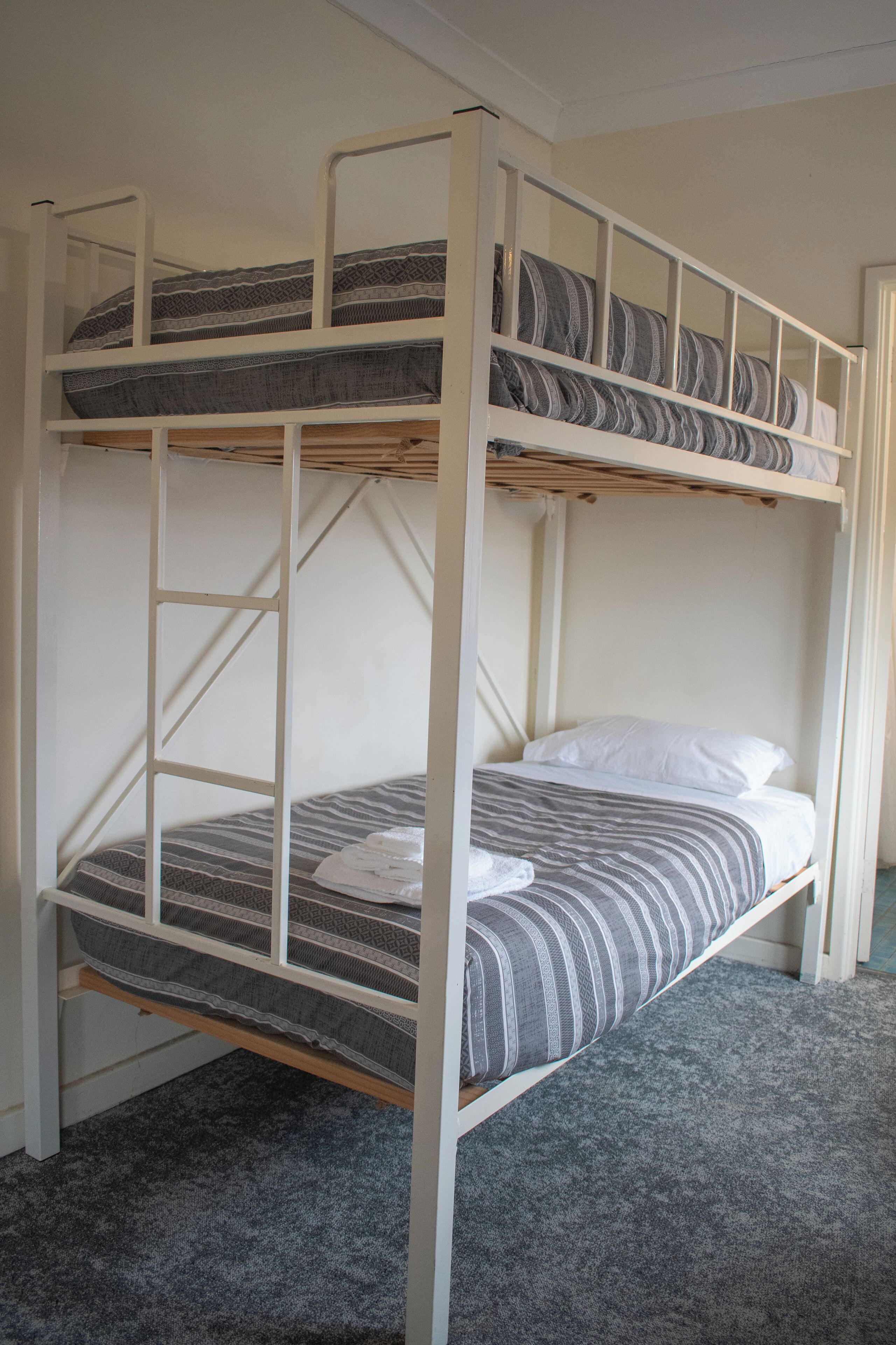 Una cama litera con escalera, ropa de cama a rayas grises y blancas, y un piso cubierto de moqueta gris.