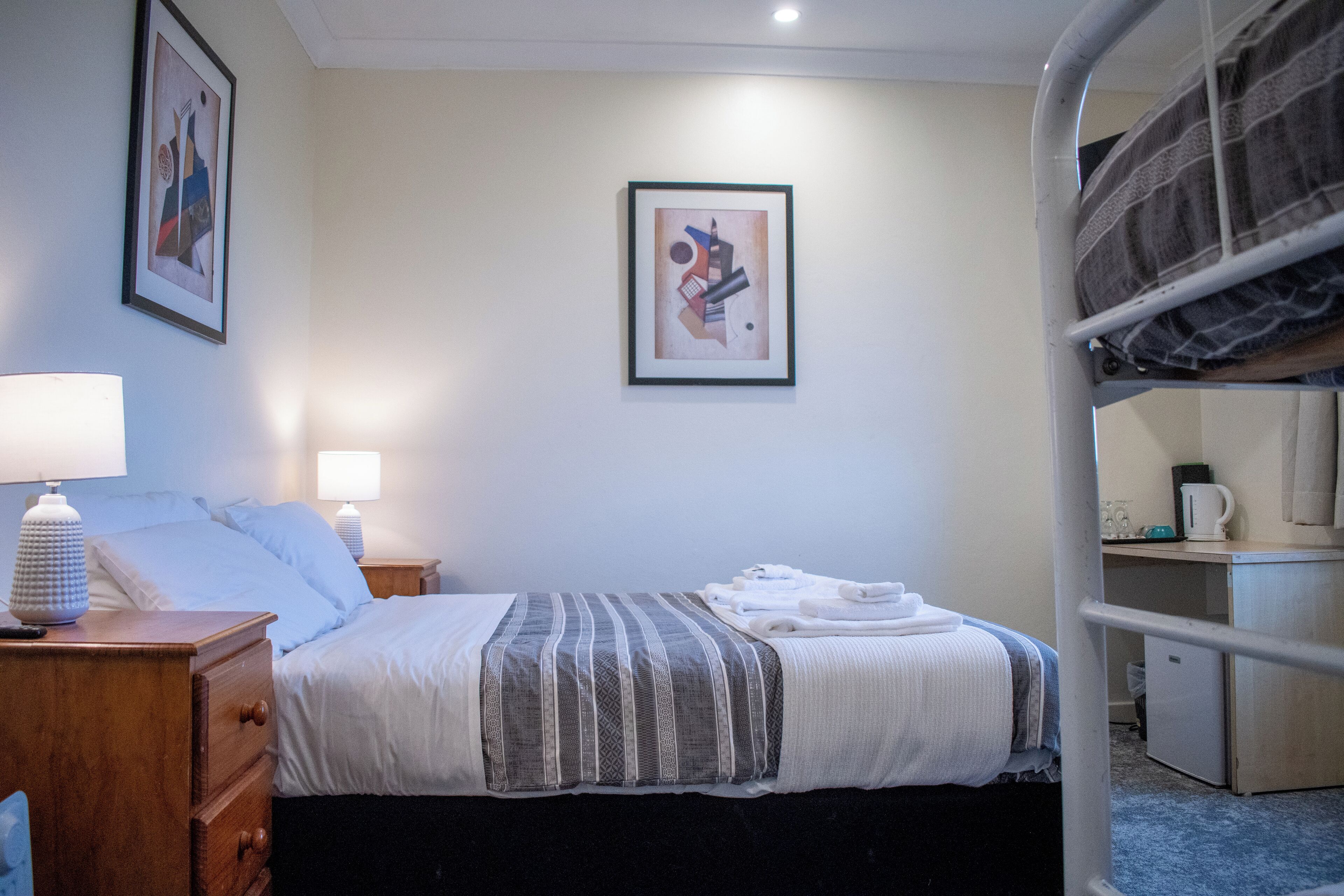 Une chambre d’hôtel avec un lit simple, une table de chevet avec une lampe, une commode, un tableau abstrait encadré accroché au mur et un lit superposé dans un coin.
