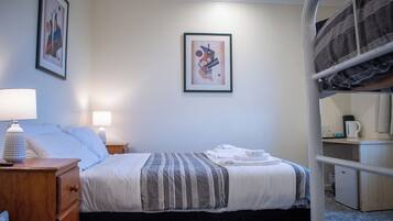 Une chambre d’hôtel avec un lit simple, une table de chevet avec une lampe, une commode, un tableau abstrait encadré accroché au mur et un lit superposé dans un coin.