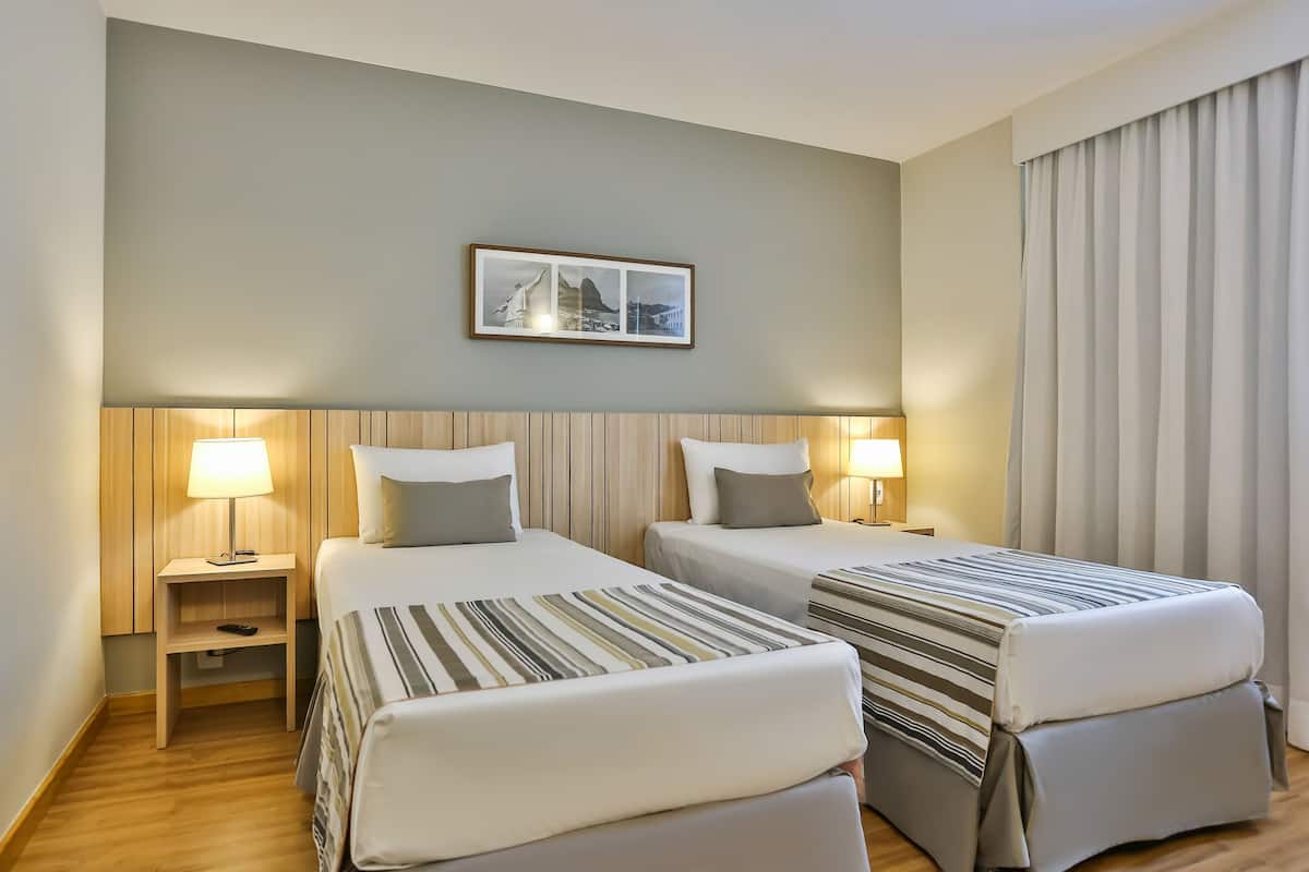 A hotel room with two beds, a nightstand with a lamp, a framed picture on the wall, and a window with curtains.