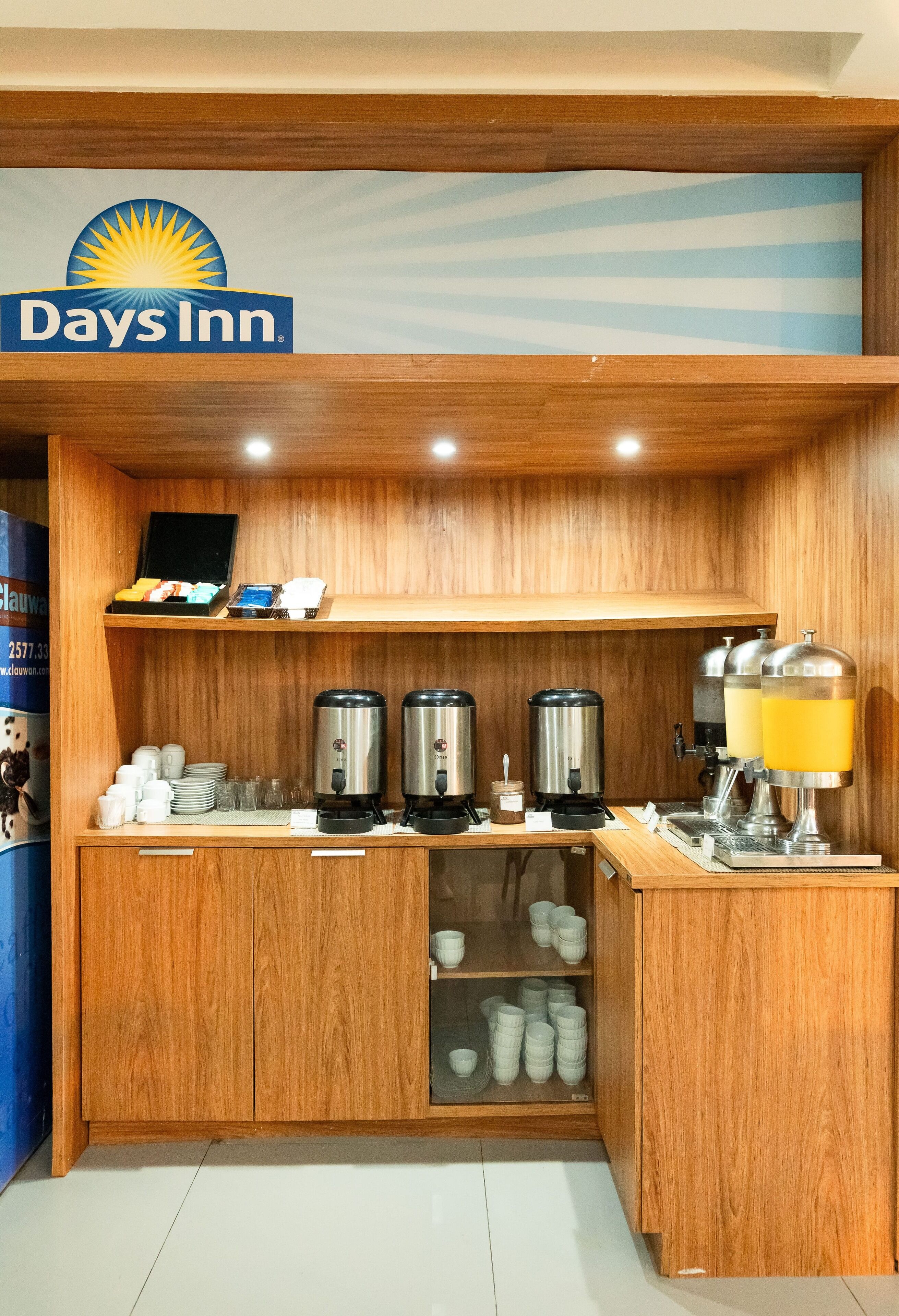 A Days Inn kitchenette with coffee makers, a juice dispenser, and a shelf with cups and mugs.