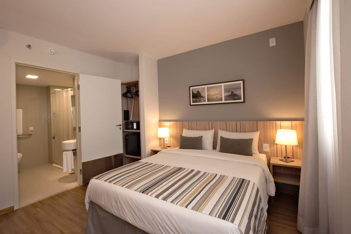 A hotel room with a bed, two bedside lamps, a nightstand, a small table, a wall-mounted picture, and a bathroom with a shower and toilet.