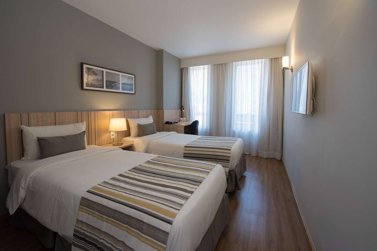 A hotel room with two beds, a nightstand with a lamp, a desk, a chair, a window with curtains, and a framed picture on the wall.
