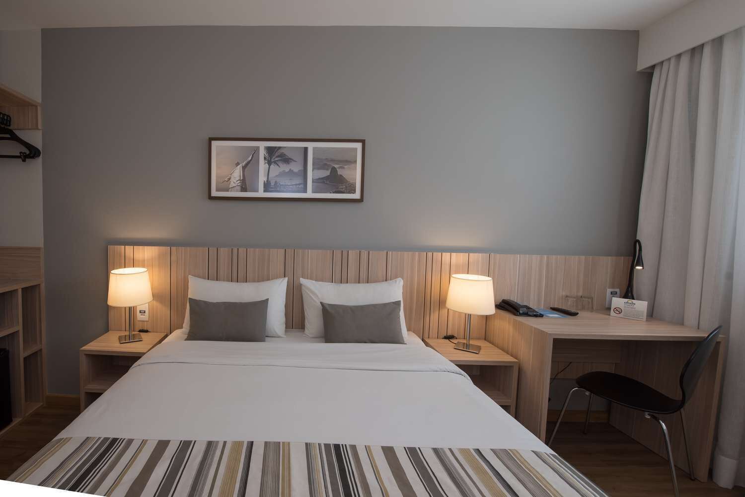 Ein Hotelzimmer mit einem großen Bett, zwei Nachttischen, einem Schreibtisch und einem Stuhl. Über dem Bett hängt ein gerahmtes Bild an der Wand.