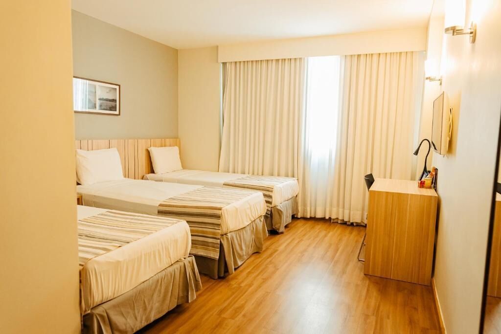 Habitació d'hotel amb dos llits, un terra de fusta, un taulell petit i una finestra amb cortines.