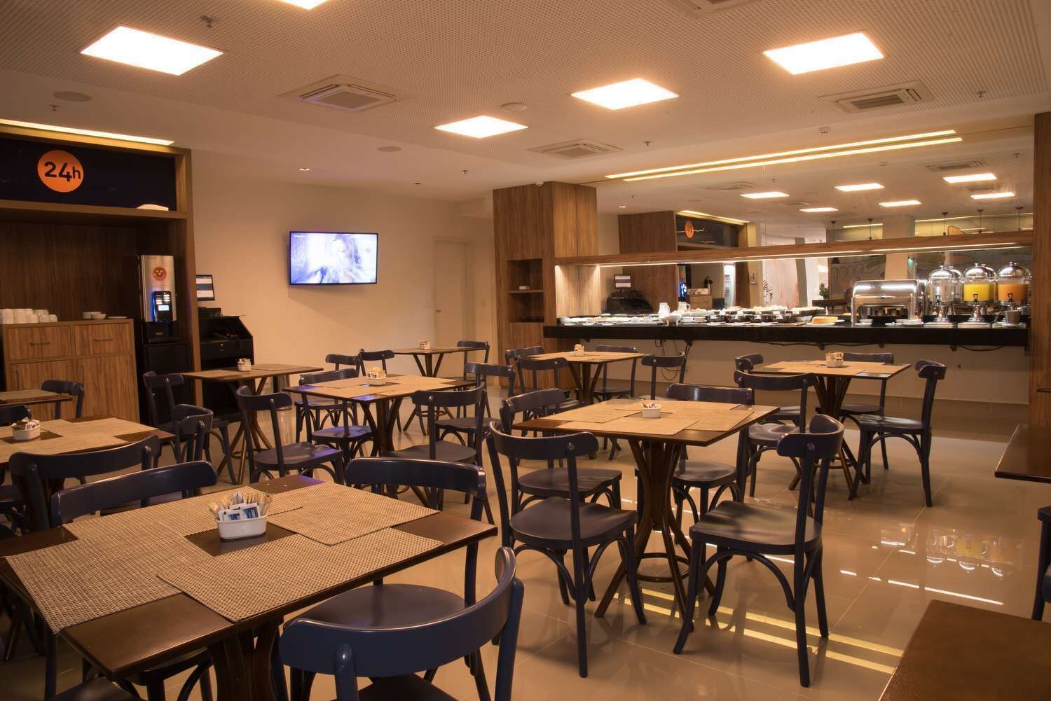 Un restaurante con varias mesas y sillas, un televisor montado en la pared y un mostrador de buffet al fondo.