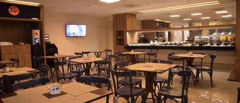 Un restaurante con varias mesas y sillas, un televisor montado en la pared y un mostrador de buffet al fondo.