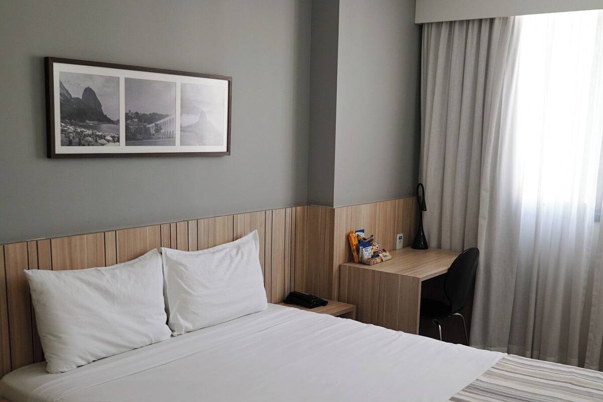 A hotel room with a large bed, a desk, a chair, a lamp, and a framed picture on the wall.