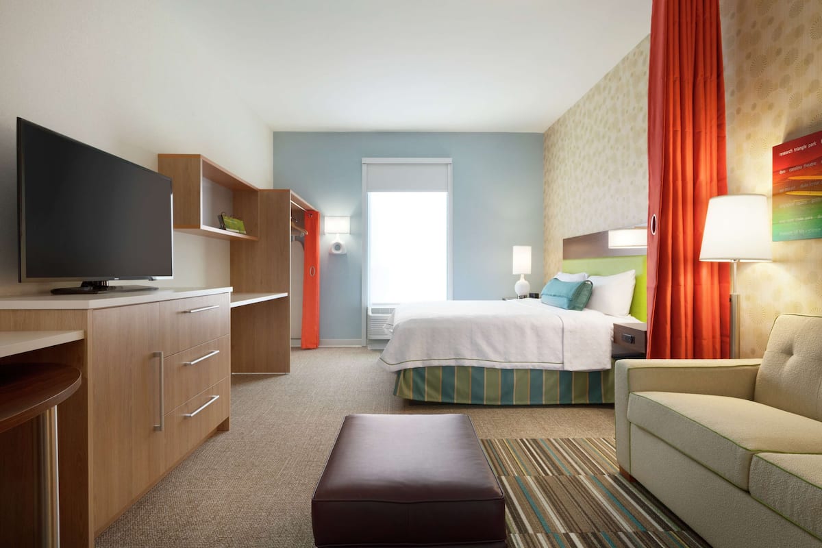 A hotel room with a television, a bed with a green and white striped bedspread, a sofa, a desk with drawers, and a window with curtains.