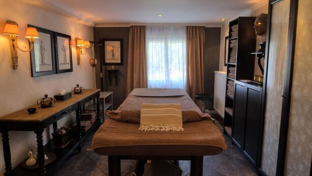 A room with a massage table, a wooden console table with tea sets, a bookshelf with towels, and a large window with curtains. - Landhotel Kern (Bad Zwesten)