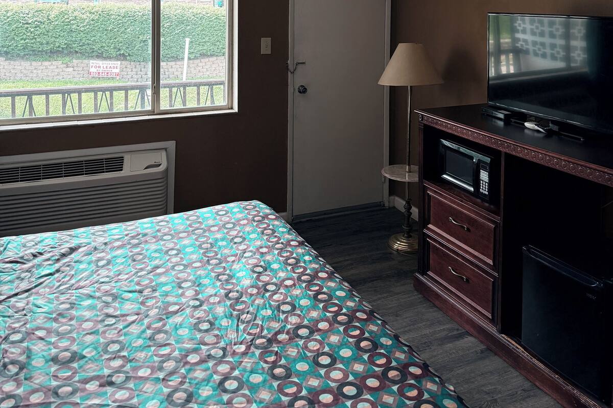 A hotel room with a bed, a television, a microwave, and a window with a view of a building and greenery outside.