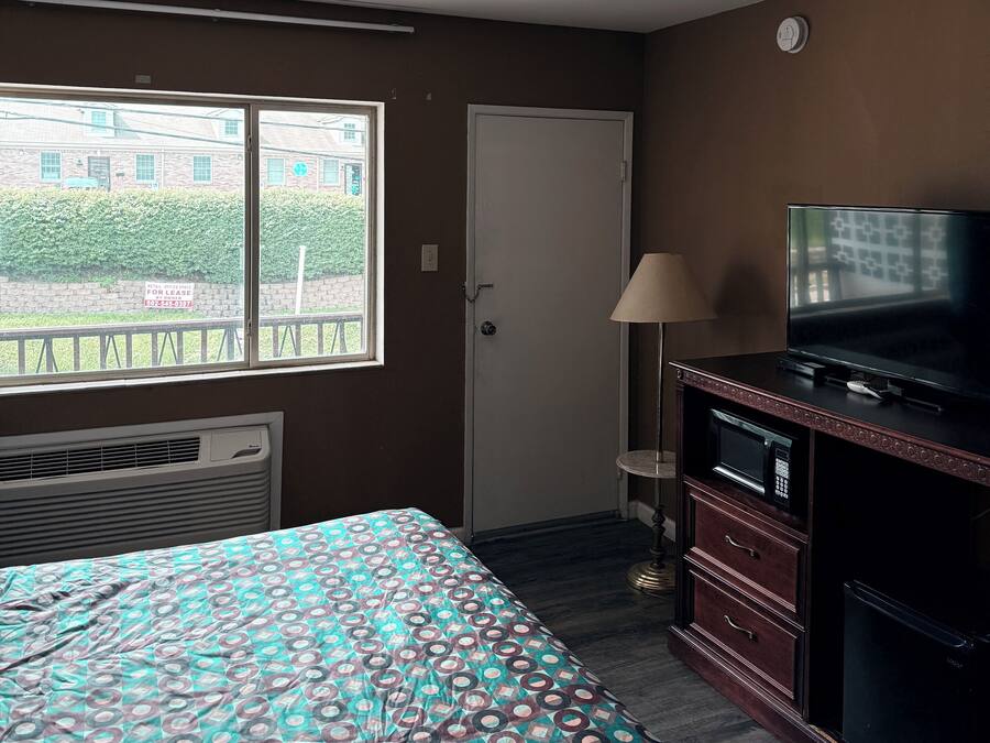 A hotel room with a bed, a television, a microwave, and a window with a view of a building and greenery outside.