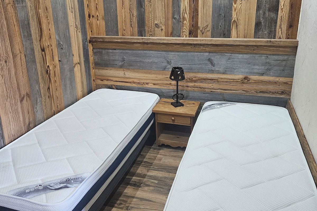 A room with two beds, wooden walls, and a nightstand with a lamp.