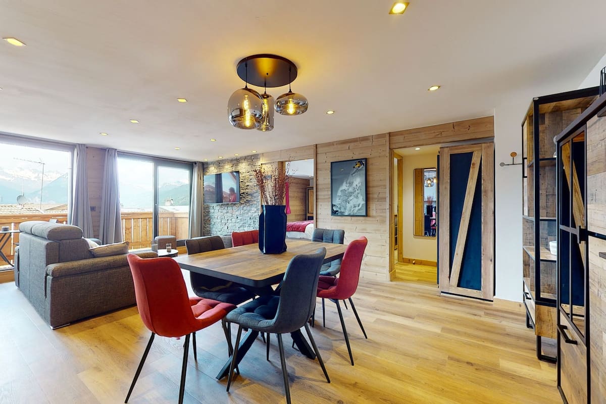 A modern dining area with a wooden table and chairs, a stone wall, and a view of mountains.