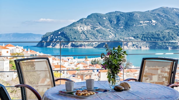 Una mesa con vistas al mar y a las montañas, dos sillas, un florero con flores y un plato de comida.
